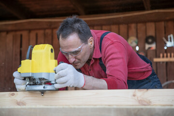 Handsome middle-aged carpenter works on wooden workpiece with a milling tool. Processing of Natural Wooden beam with milling woodworking machine. Working with hand tool fraser on wood