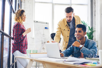 Obraz premium Positive male and female staff members watching funny video on laptop computer connected to internet and laughing while preparing creative task,young male having funny messaging in social network