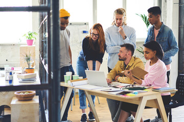 Mature male leader of diversity working group of graphic designers supervising work of staff taking project creation on laptop computer giving advice using teambuilding concept for collaborating