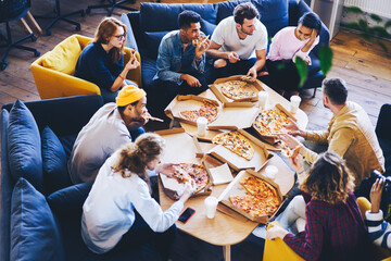 Fototapeta na wymiar Group of male and female coworkers having lunch break eating pizza together in office, overhead shot of young casually dressed staff members recreating after finishing tasks making fast snack