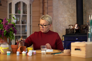 middle-aged woman choosing pencil to draw in cozy country house with fireplace Concentrated woman with blond hair dressed casually painting