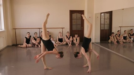 Two flexible young ballerinas are standing in the vertical splits in a ballet class, with ballerinas sitting in the background. A group of young ballerinas rehearsing in a dance Studio.