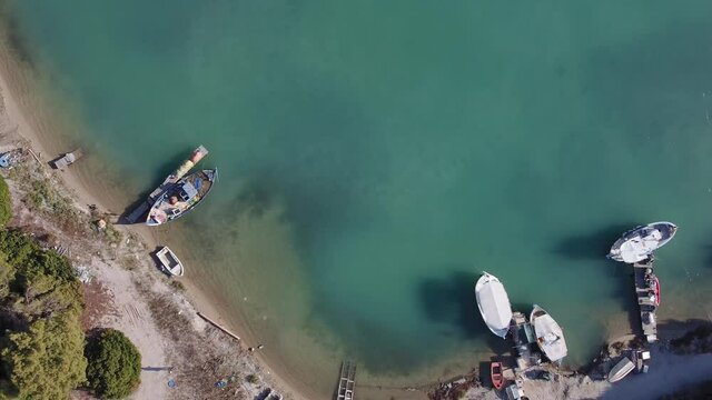 Coastal drone top view of moored boats on lagoon with green plantation. Aerial shot of caiques, fishing and modern small ships at Glarokavos in Kassandra Chalkidiki peninsula with calm waters.