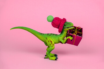 cute green plastic toy dinosaur wearing knitted hat and holding present box  pink background