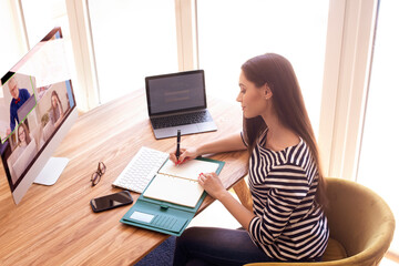 High angle shot of young woman writing something into her notebook while sitting at desk and working