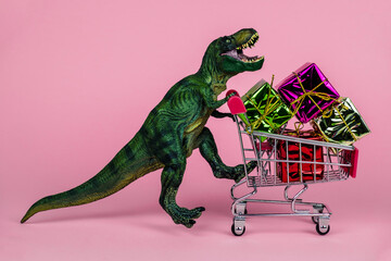 funny green dinosaur toy with shopping cart full of present boxes on a soft pink background