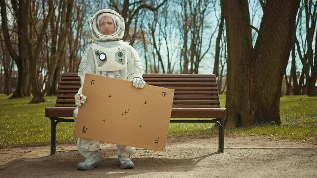 Sad Man in Spacesuit is Sitting on a Bench in a Park and Holding a Carboard Mockup Sign. Miserable Astronaut Looks in the Distance. Emotionally Depressed Spaceman in White Futuristic Suit.