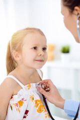 Doctor examining a child by stethoscope in sunny clinic. Happy smiling girl patient dressed in bright color dress is at usual medical inspection