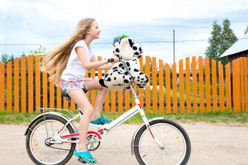 A beautiful caucasian girl with long blond fluttering hair rides a white bicycle along an orange fence. The girl carries a toy dog ​​on a bicycle. Digital detox, no gadgets, sports, relaxation.