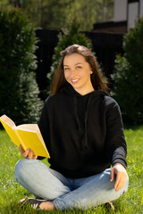 Portrait of Attractive smiling long hair brunette girl dressed in black hoodie sitting on grass...