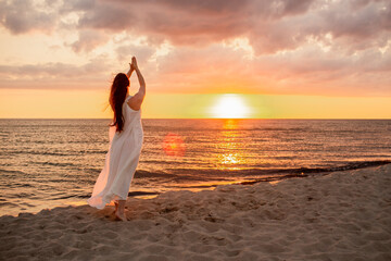 Happy young woman in a long white dress looking at the sunset on empty sand beach with her hands...