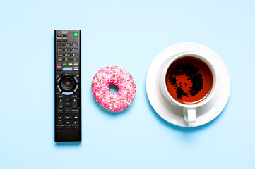 Black TV, audio remote control, cup of tea, pink sweet donut on blue background flat lay top view copy space. Minimalistic background with remote control. Concept of spending time in front of the TV