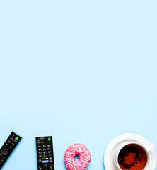 Black TV, audio remote control, cup of tea, pink sweet donut on blue background flat lay top view copy space. Minimalistic background with remote control. Concept of spending time in front of the TV