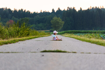 Baby sitting in the middle of a rural road leading to forest.