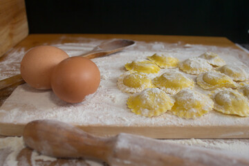 Homemade Ravioli / Tortellini /dumplings, preparing, cooking with two eggs , wooden spoon and rolling pin,on floured wood.Close up view of homemade pasta concept.Black background.