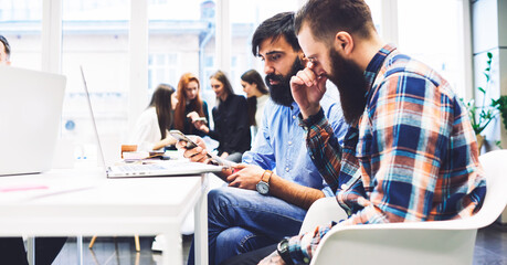Business male colleagues learning via marketing webinar using wireless connection via modern smartphone sitting in workshop.Innovative bloggers thinking over the internet content while watching videos