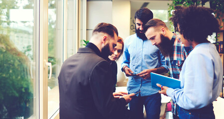 Obraz premium Team of young male and female freelancers sharing multimedia files downloading them from web page via modern smartphone connected to wireless fast 4G internet on informal meeting in coworking office