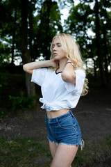 Closeup portrait of a sensual blonde curly young woman with closed eyes holding her hands behind neck, dress denim shorts walking in park.
