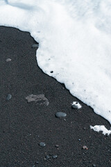 Oceanic wave with white foam rolls over black sand beach with pebble . Silky black beach texture. Minimalistic black background. Tenerife voulcanic sandy shore.