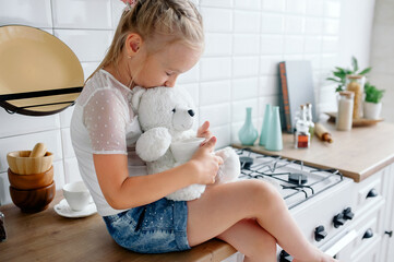 baby girl hugs a teddy bear sitting in a beautiful stylish bright kitchen. Teddy Bear Day September...