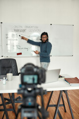 Live stream. Young bearded male teacher wearing glasses pointing at whiteboard and giving...