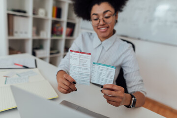 Happy afro american female teacher sitting at her workplace and showing paper cards with rules of English grammar at webcam. Working from home