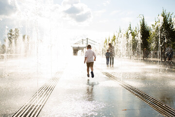 Unknown little boy in a white shirt runs his back among the fountains in the square in the summer on a hot day