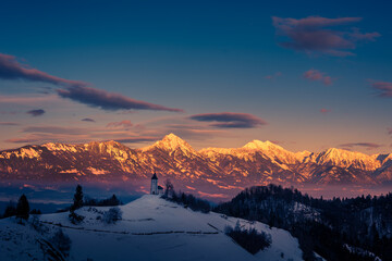 Fototapeta na wymiar Gorgeous and colorful sunrise over Jamnik church with Alps mountains range in the distance. Famous landmark in Slovenia in wintertime. Landscape covered with snow