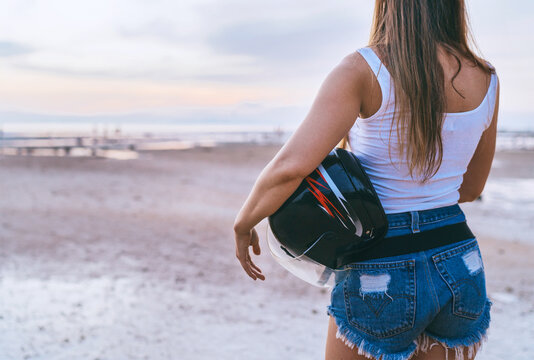 Young long haired woman dressed sexy jeans shorts making moto trip rest with motorcycle helmet enjoying a sunset sky on Koh Samui island, Thailand. Traveling in exotic countries concept image.