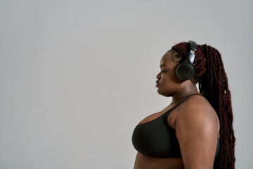 Fresh start. Plump, plus size african american woman in sportswear and headphones listening to music, standing in studio over grey background. Concept of sport, healthy lifestyle, body positive