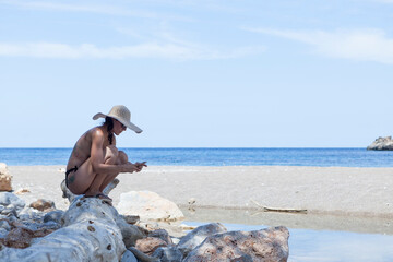 A pretty young  woman on a lonely Mediterranean sea beach in Majorca. The woman is wearing a black bikini and a straw hat. The woman is taking photos and selfies with her mobile phone on the beach.