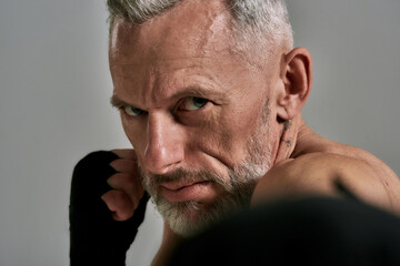 Close up of middle aged athletic man, kickboxer looking agressive while boxing, training in studio over grey background. Muay Thai, Boxing or Kickboxing concept