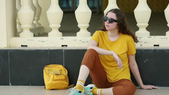 A teenage girl in a yellow t-shirt, brown breeches and sunglasses with a yellow backpack sits at the decorative fence of a city Park