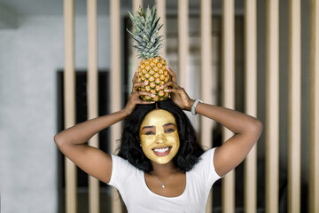 Fresh summer shot of joyful African girl in white t-shirt with pineapple on head, golden peel off mask on her face, looks at camera and smiles broadly, standing on cozy home room background