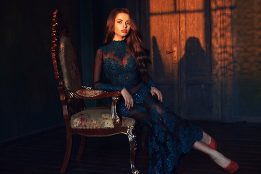 Fine art photo of young elegant beautiful lady wearing long green evening dress with lace and rhinestones and sitting in armchair in interior in sunset lightning.