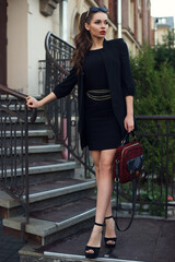 Young beautiful stylish classy girl wearing black dress and coat, holding red handbag, standing and...
