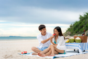 Asian young couple sitting on the picnic blanket and eating watermelon in the beach and near sea with tropical fruit in background. Summer, holidays, vacation and happy people concept