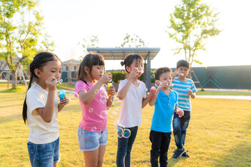 Large group of happy Asian smiling kindergarten kids friends playing blowing bubbles together in the park on the green grass on sunny summer day.