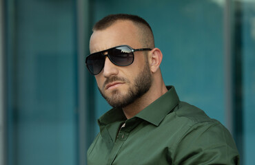 Obraz premium Confident handsome man in sunglasses and shirt over city background. Men's beauty, fashion. Outdoor portrait.