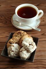 Oatmeal cookies with walnuts and raisins, meringue cookies with hazelnuts in a square bowl and a cup of black tea on a wooden table. Closeup