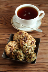 Oatmeal cookies with walnuts and raisins, sugar-coated pumpkin seed cookies in a square bowl and a cup of black tea on a wooden table. Closeup