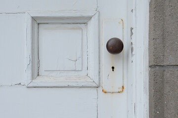 A vintage white wooden door with a brown metal knob and small keyhole.There's a square panel in the door adjacent to the door knob. The old door is peeling and is attached to a brick wall.