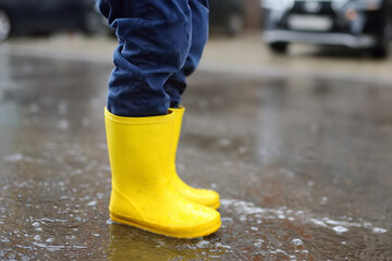 Little boy wearing yellow rubber boots jumping in puddle of water on rainy summer day in small town. Child having fun.