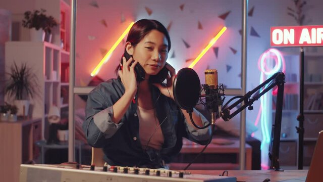 Happy young Asian woman taking off headphones, looking at camera and smiling while sitting at desk in home radio studio with colorful neon light and on-air sign