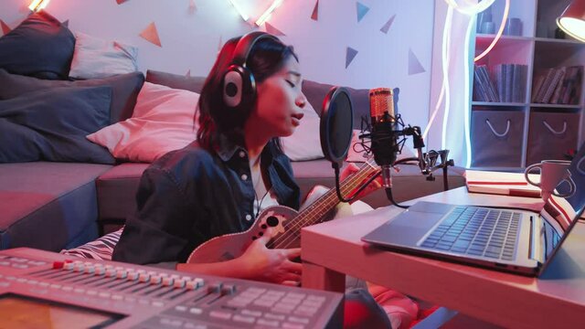 Young Asian female musician in headphones sitting on the floor in home recording studio with neon lamps, playing the guitar and singing a song in front of microphone
