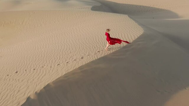 Nature and travel 4K aerial video. A young female in a beautiful fluttering red dress is in the desert landscape. Drone footage of a woman making footprints on the sand dunes at sunset on a windy day