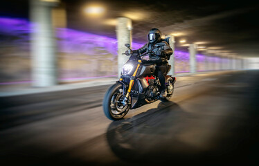 Motorcyclist in full black leathers and helmet riding fast through tunnel with columns and purple light in background