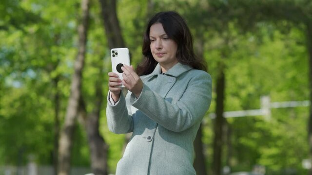 Beautiful adult woman taking picture by mobile phone in park brunette woman holding modern smartphone photographing on sunny spring day.