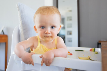 Adorable blue eyed baby girl with green puree smudges on face sitting in highchair and looking at camera. Closeup shot. Feeding process or child care concept