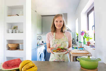 Happy woman cooking at home, keeping healthy diet, holding homemade vegetable dish bowl, smiling at...